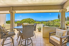 An outdoor patio with wicker furniture, a round table, and a view of a lush green landscape. - FRESH Competitive Pricing, (16T-8), Parking Level, Easy Access, VERY NICE ~ Platinum Maui Vacations (Lahaina)
