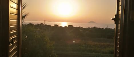 An open window with a view of a sunset over a body of water, with land and vegetation in the foreground.