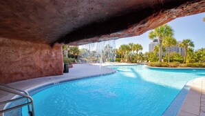 Ein Swimmingpool unter einem natürlichen Felsbogen, umgeben von Palmen und einem klaren Himmel.