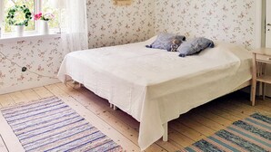 Un dormitori amb llit, almoines, una tauleta de nit, una finestra amb cortines, un paper pintat amb motius florals i un quadre petit a la paret.
