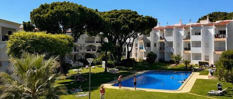A pool area with a clear blue sky, surrounded by well-maintained grass, palm trees, and a building with multiple balconies.