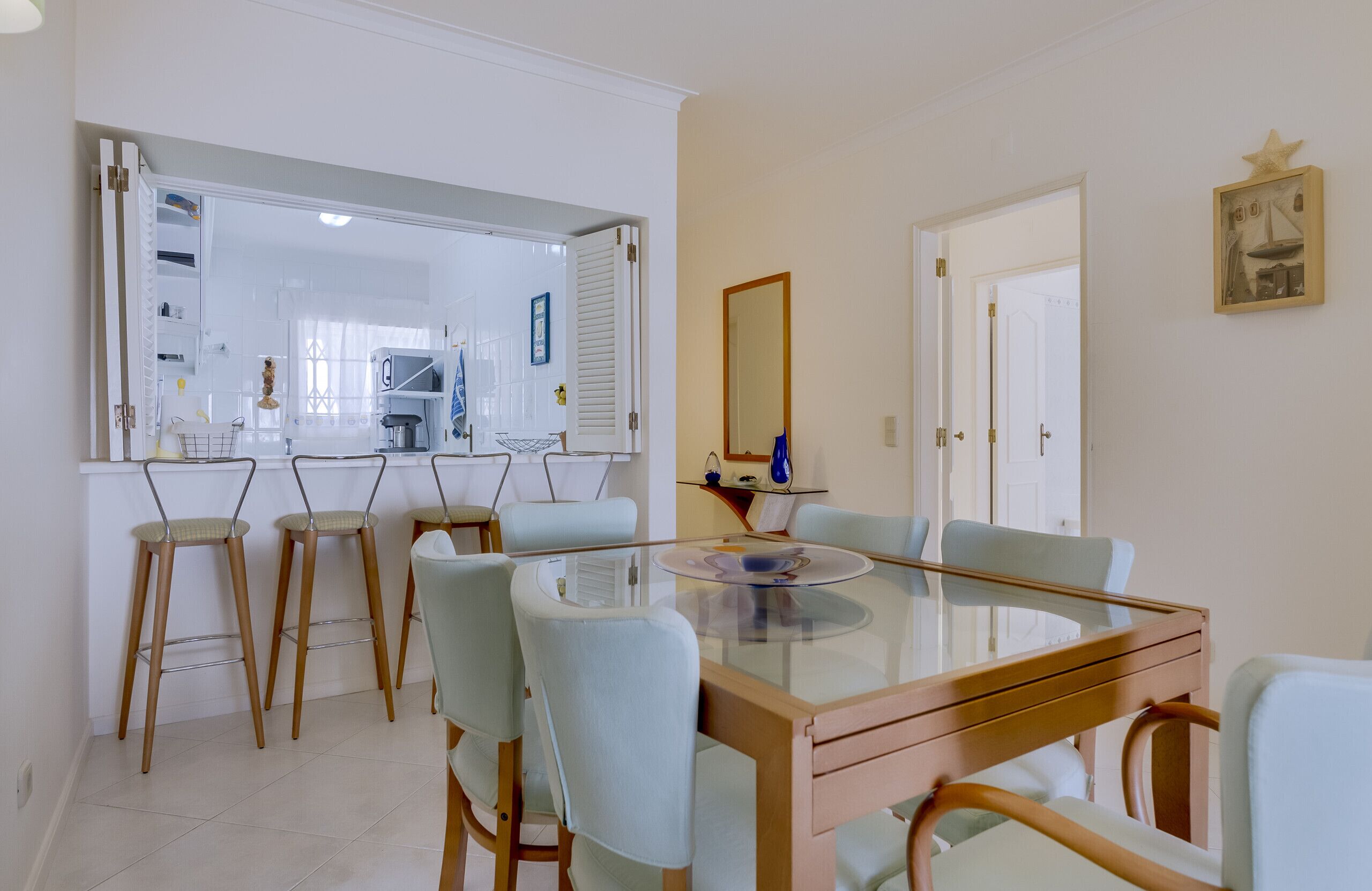 A dining area with a glass-top table and four chairs, a kitchen in the background, and a door leading to another room.