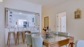 Un coin repas avec une table à plateau de verre et quatre chaises, une cuisine en arrière-plan et une porte donnant sur une autre pièce.