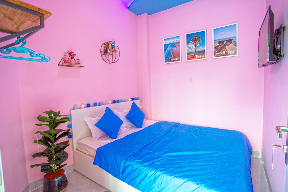 A pink room with a bed, a plant, and three framed pictures on the wall.