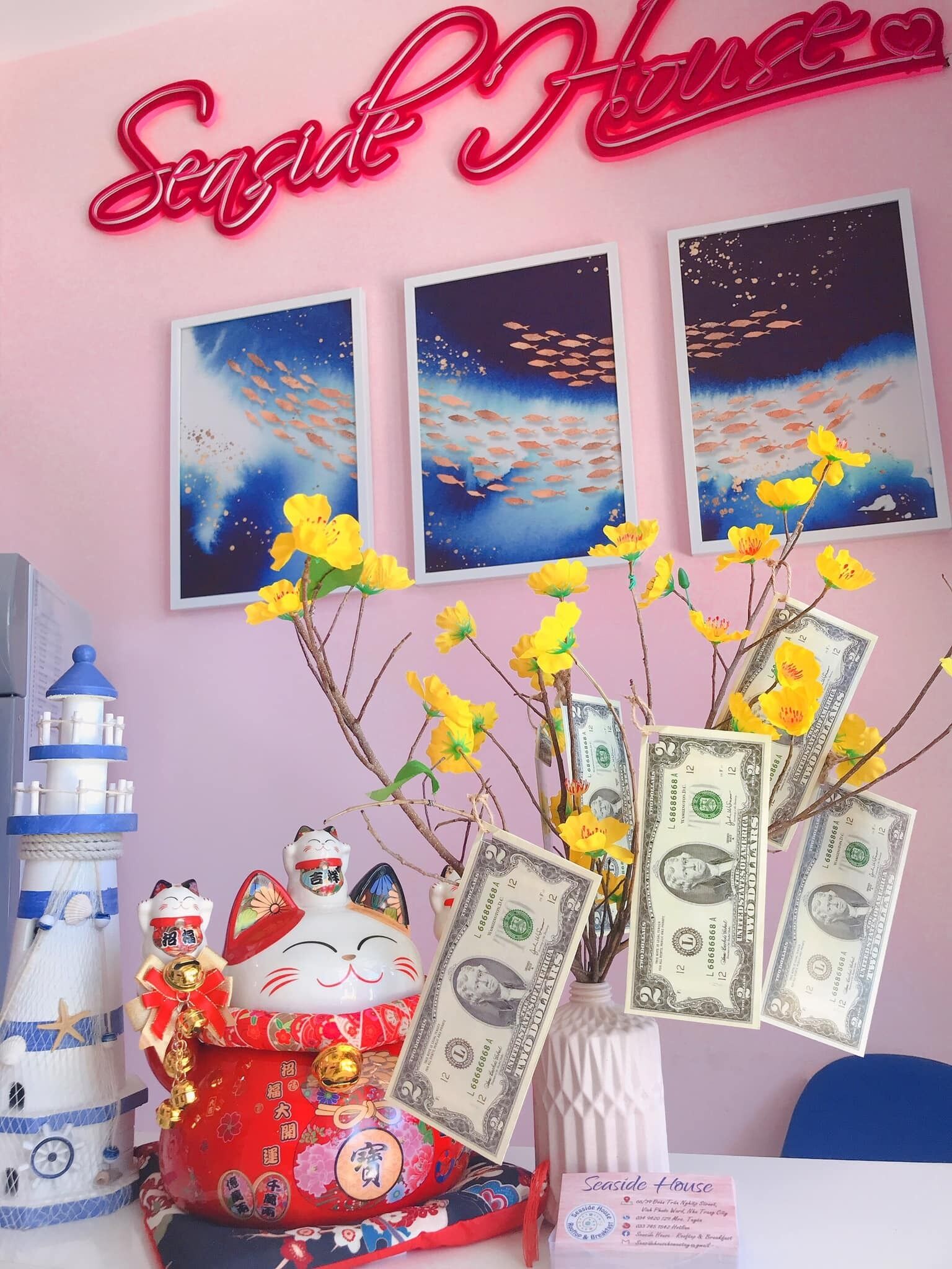 Une pièce avec un mur rose, un panneau néon indiquant « Seaside House », trois tableaux encadrés représentant des poissons, un vase décoratif avec des fleurs jaunes, un petit phare et une figurine de chat tenant un billet d’un dollar.