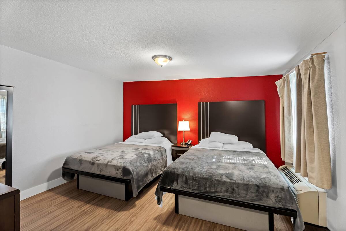 A hotel room with two beds, a red accent wall, a ceiling light, and a window with curtains.