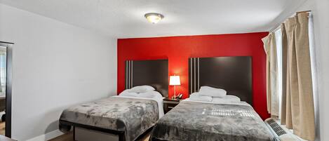 A hotel room with two beds, a red accent wall, a ceiling light, and a window with curtains.