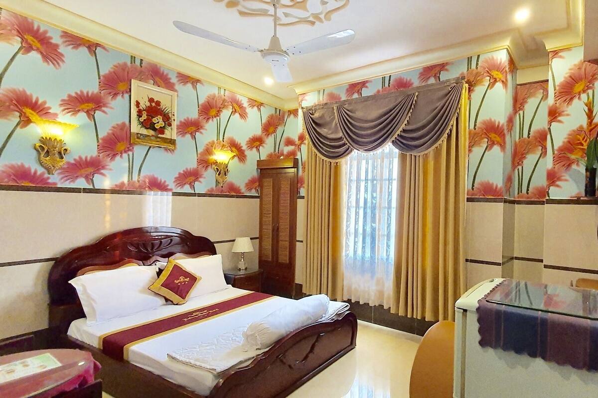 Une chambre d’hôtel comprenant un lit, une table de chevet, un bureau, une chaise, un ventilateur de plafond et un papier peint à motifs floraux.