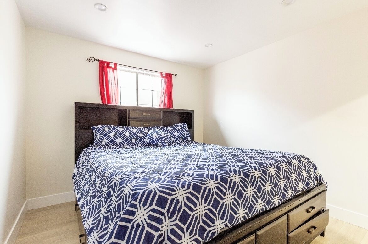 Een slaapkamer met een bed, nachtkastje, kapstok en een raam met rode gordijnen.