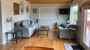 Un salon avec un plafond en bois, deux canapés gris, une table basse, un téléviseur et une fenêtre avec des stores.