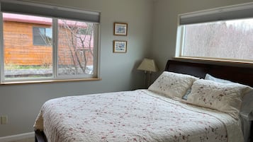 Ein Schlafzimmer mit einem Bett, zwei Fenstern, einer Lampe und zwei gerahmten Bildern an der Wand.
