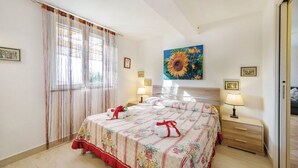 Ein Hotelzimmer mit einem großen Bett, zwei Nachttischen mit Lampen, einem Fenster mit durchsichtigen Vorhängen und einem Gemälde einer Sonnenblume an der Wand.
