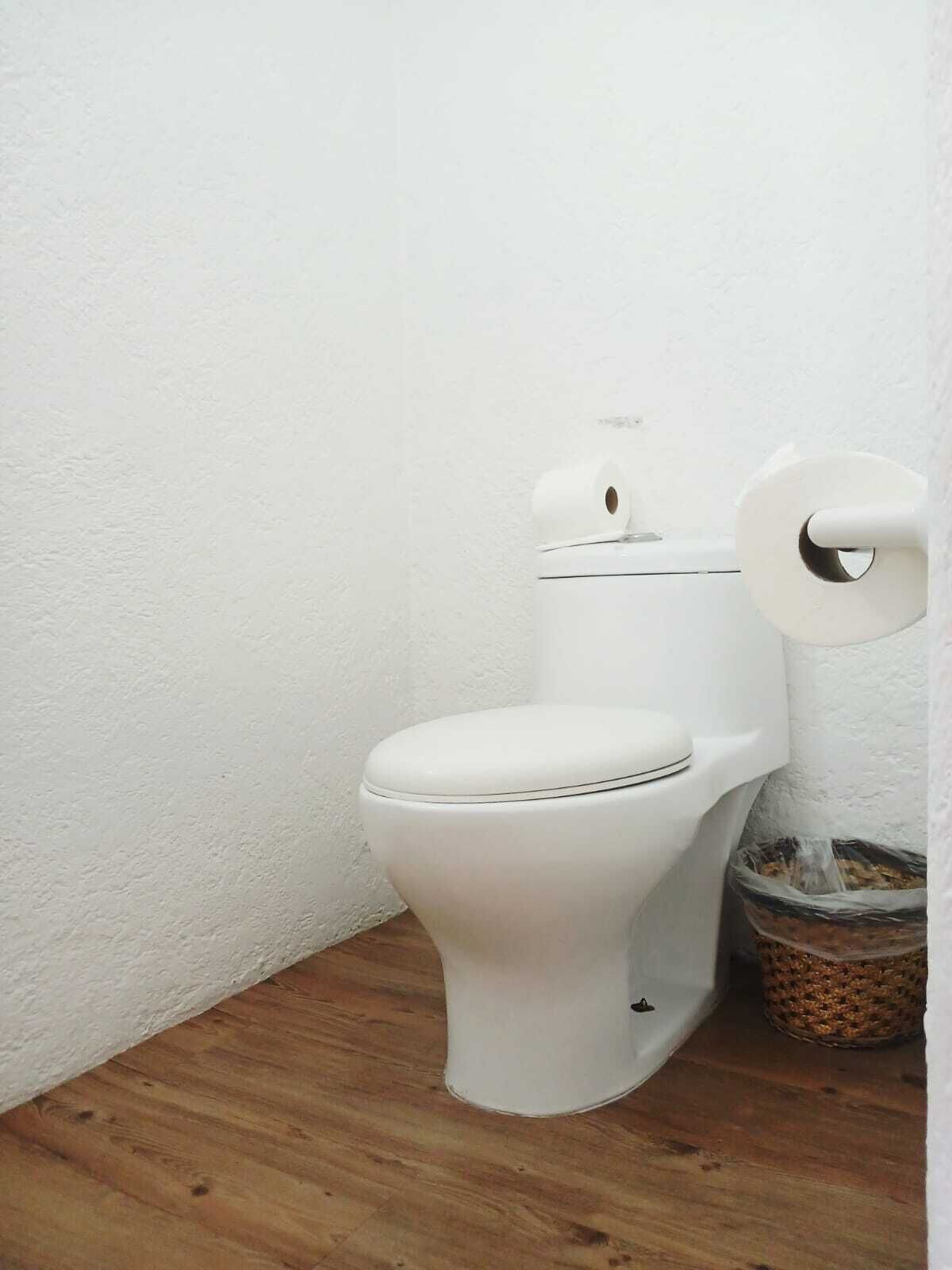A white toilet with a roll of toilet paper on the wall, a trash bin, and a wooden floor.