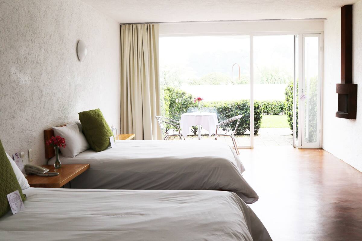 Ein Hotelzimmer mit zwei Betten, einem kleinen Tisch mit Blumenvase und Blick auf einen Garten durch eine Glastür.