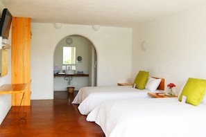 Habitación de hotel con dos camas, un escritorio de madera, una pequeña cocina y un baño visible a través de un arco.