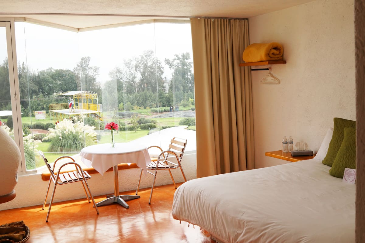 Ein Hotelzimmer mit Bett, Tisch mit Stühlen und Blick auf einen Garten, sowie einem Fenster mit Vorhängen.