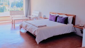 A hotel room with a bed, a chair, a small table, and a window with a view of trees.