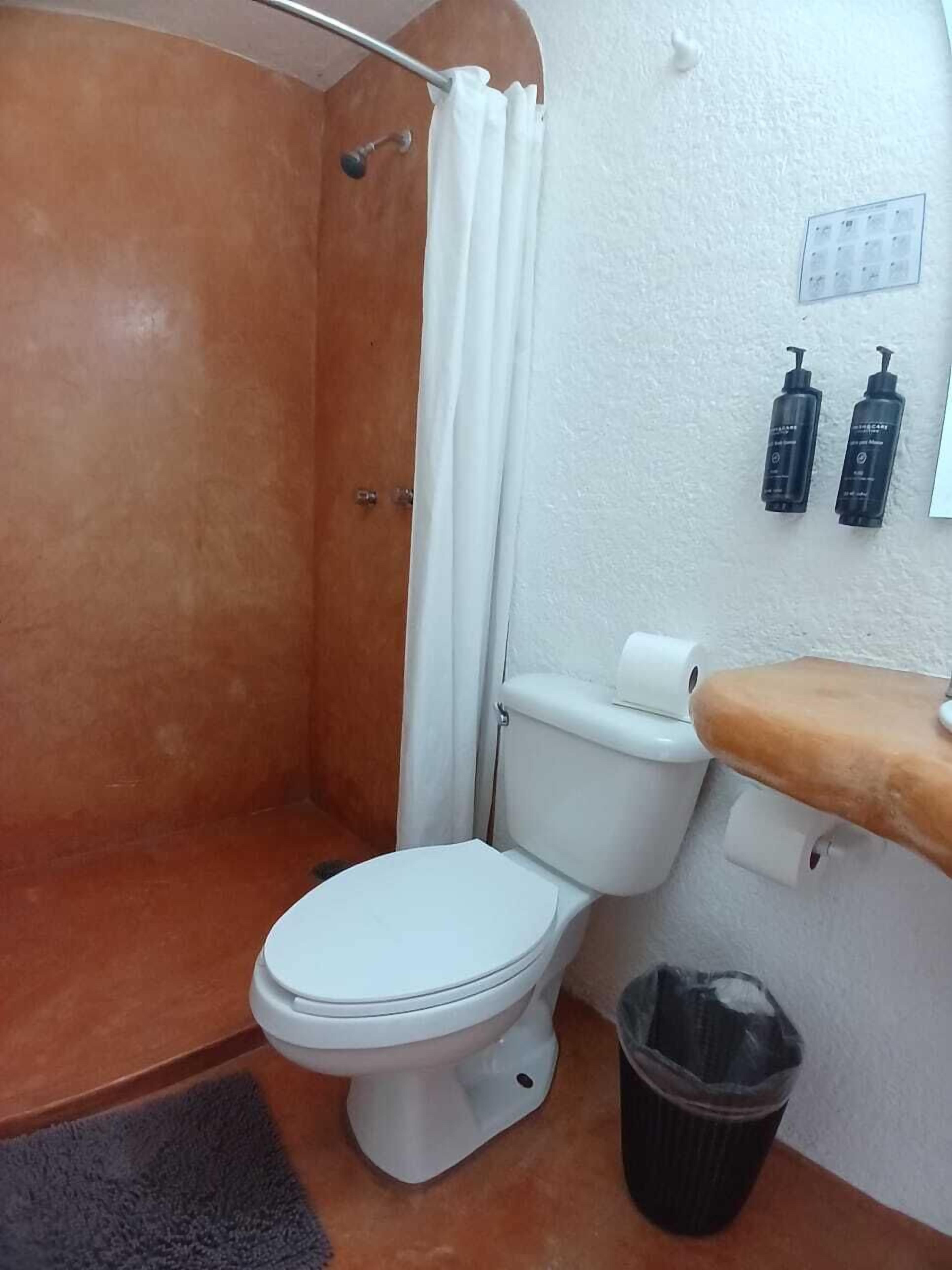 Een badkamer met een wit toilet, een douche met gordijn, een prullenbak en twee flessen aan de muur.