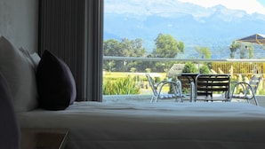 A hotel room with a bed, a view of mountains, and a balcony with chairs.