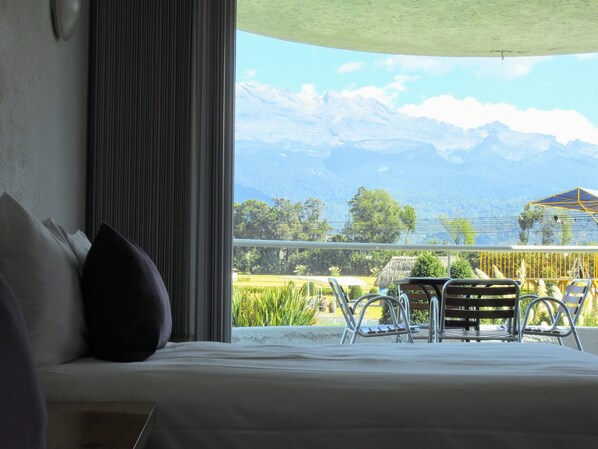 A hotel room with a bed, a view of mountains, and a balcony with chairs.