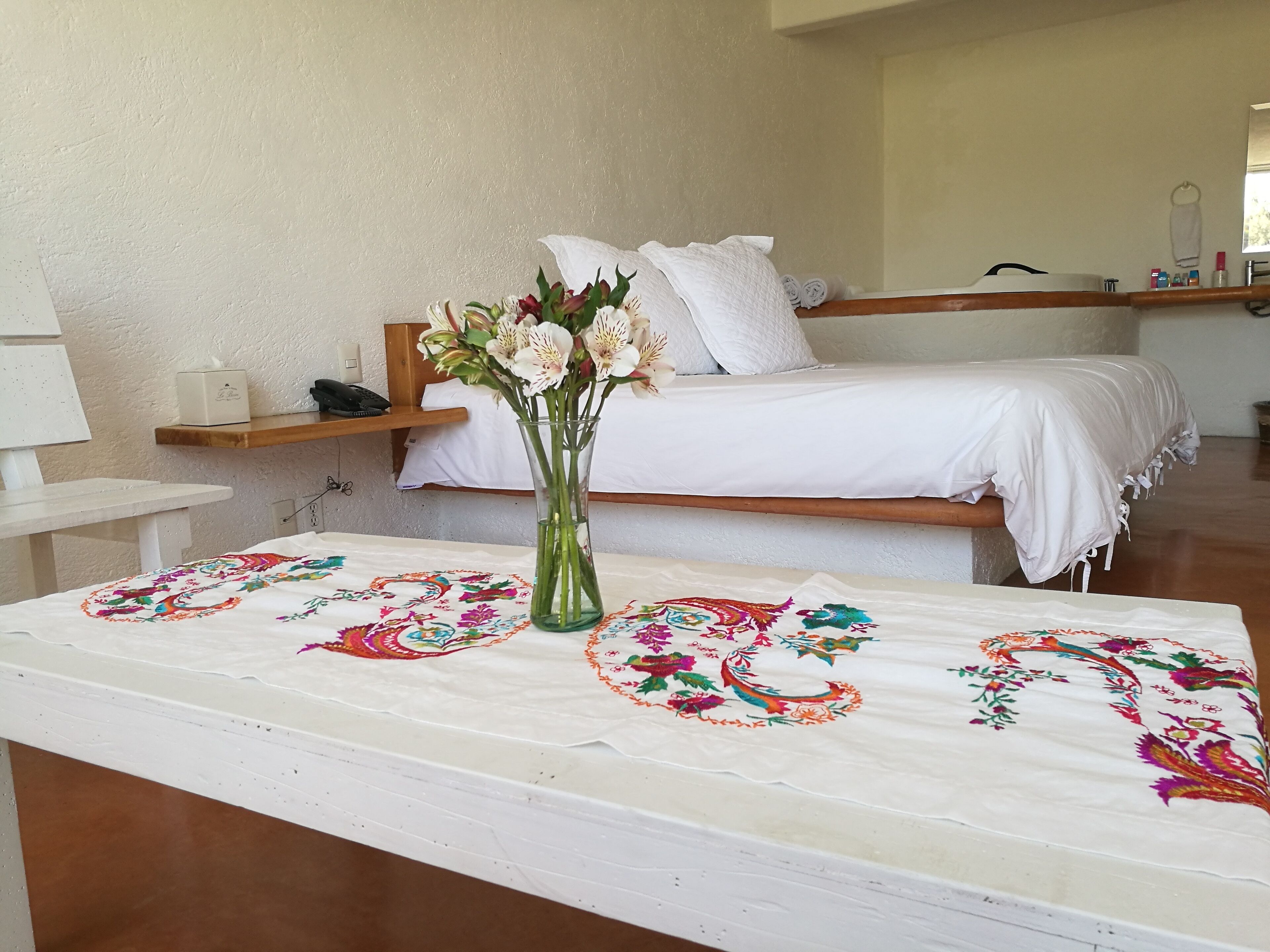 A hotel room with a bed, a bedside table, a nightstand, and a vase with flowers.