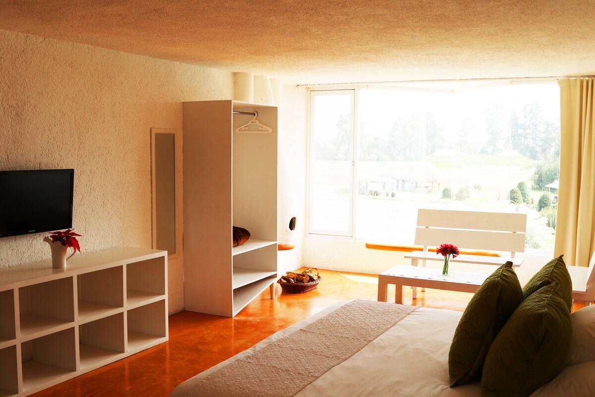 Ein modernes Hotelzimmer mit einem Flachbildfernseher, einer weißen Regaleinheit, einem Esstisch mit Blumen und einem großen Fenster mit Blick ins Freie.