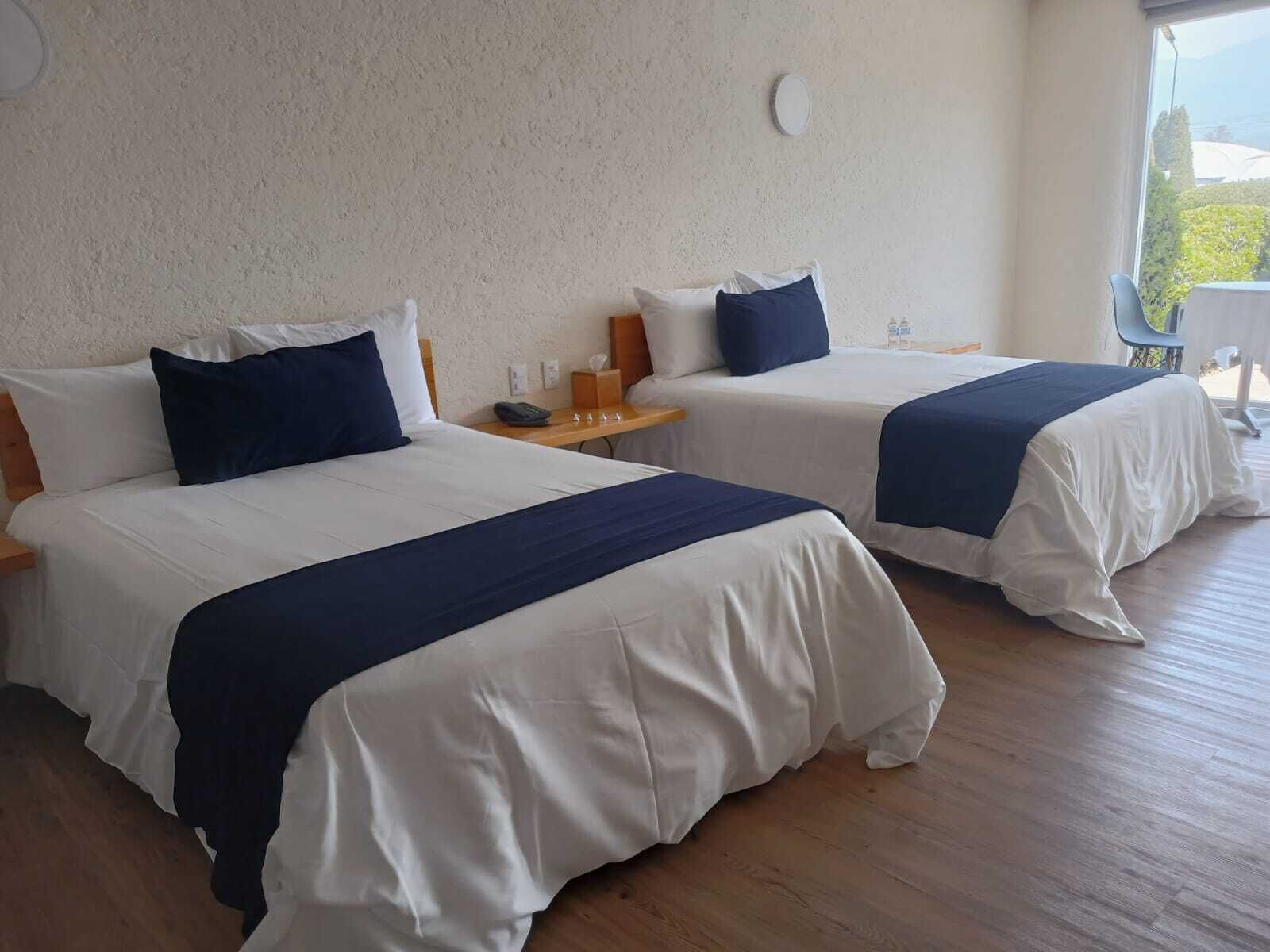 Two beds with white and navy bedding, wooden headboards, and a small table with a phone and a bottle.