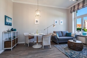 Un salon moderne avec une table à manger ronde, des chaises en osier, un canapé gris et une grande fenêtre donnant sur des immeubles.