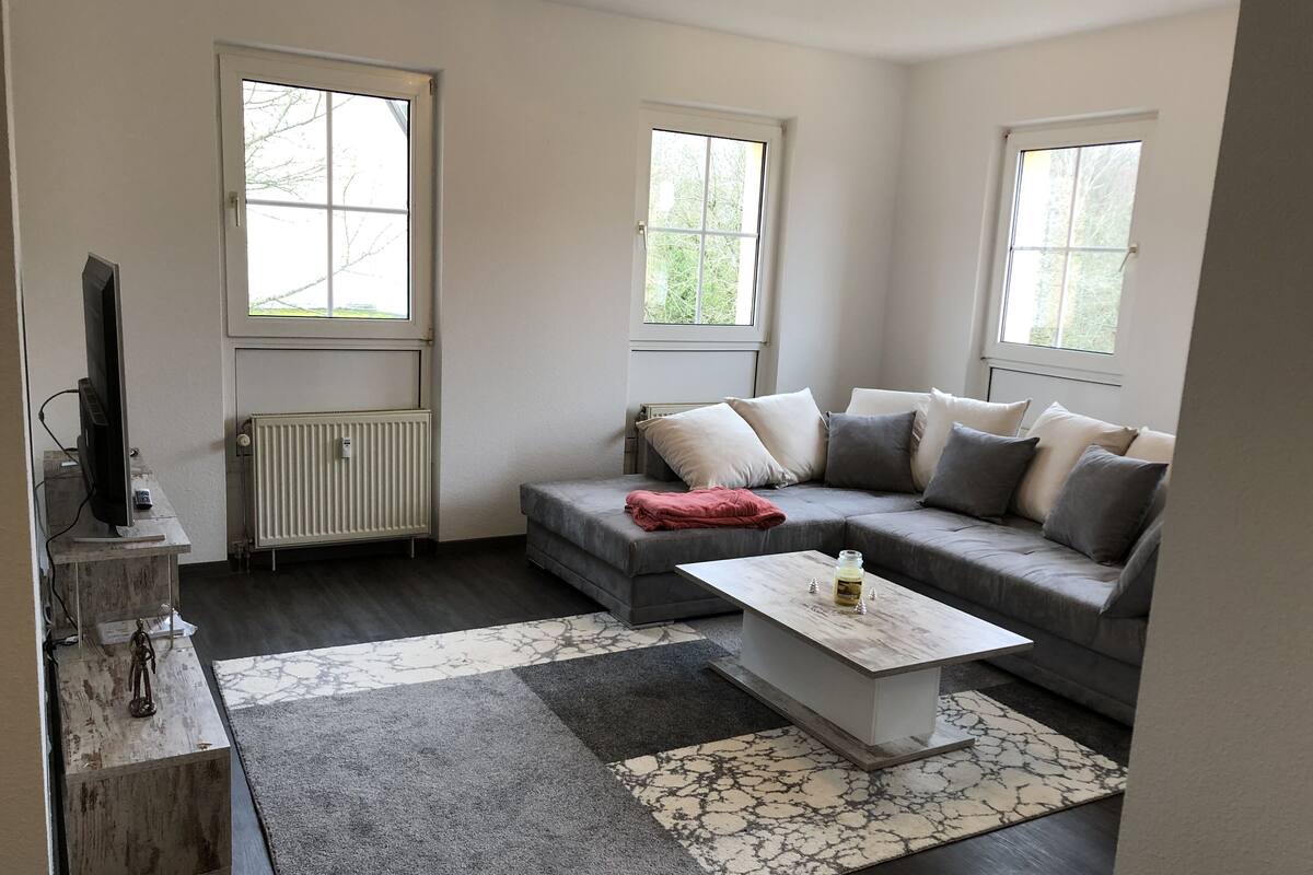 A modern living room with a grey sectional sofa, a white coffee table, and a large window providing natural light.