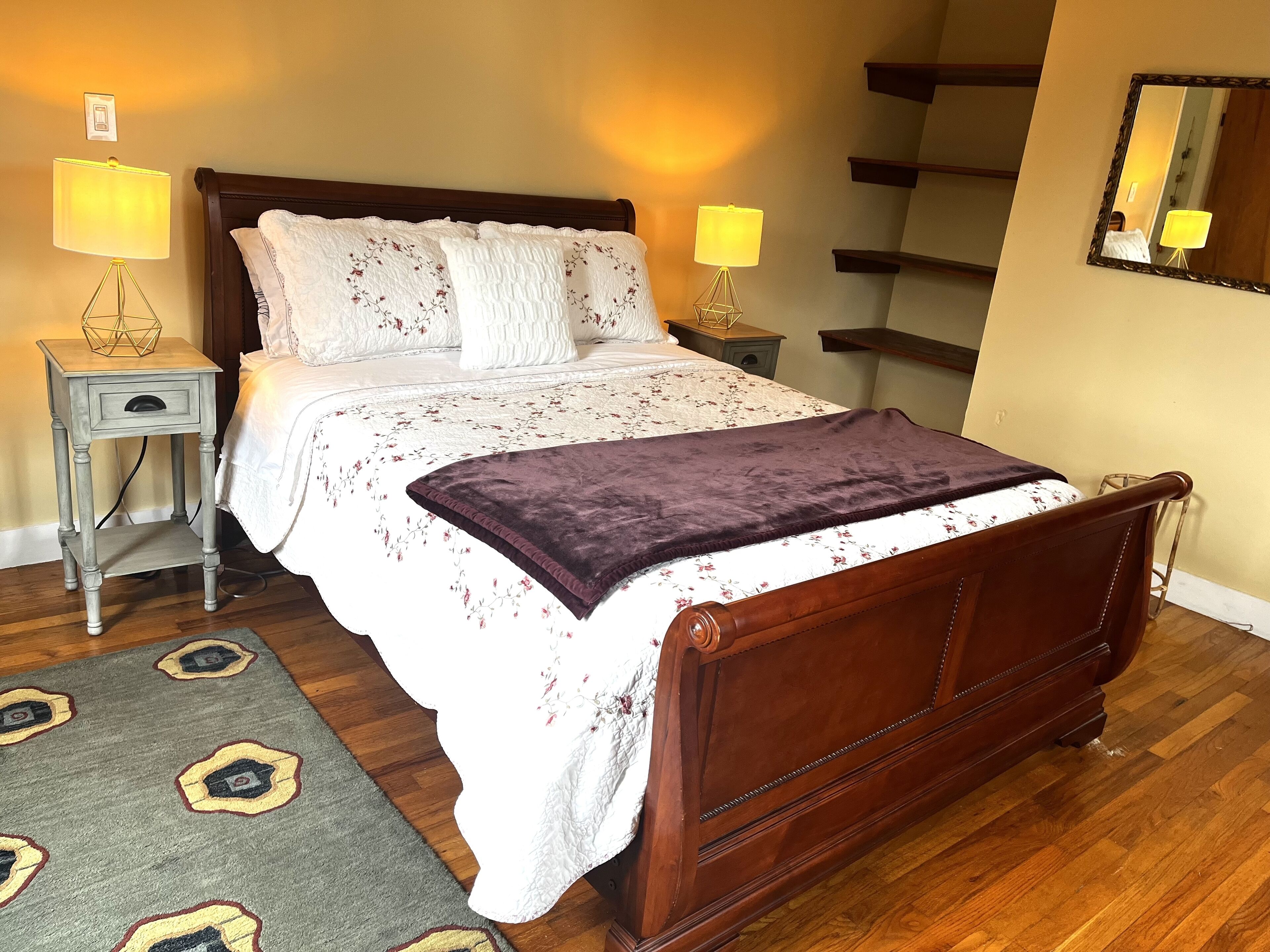 Een netjes opgemaakt bed met een bloemenbedekker en een paars plaid, met aan beide zijden nachtkastjes met lampen, in een kamer met houten vloeren en een grijs vloerkleed.