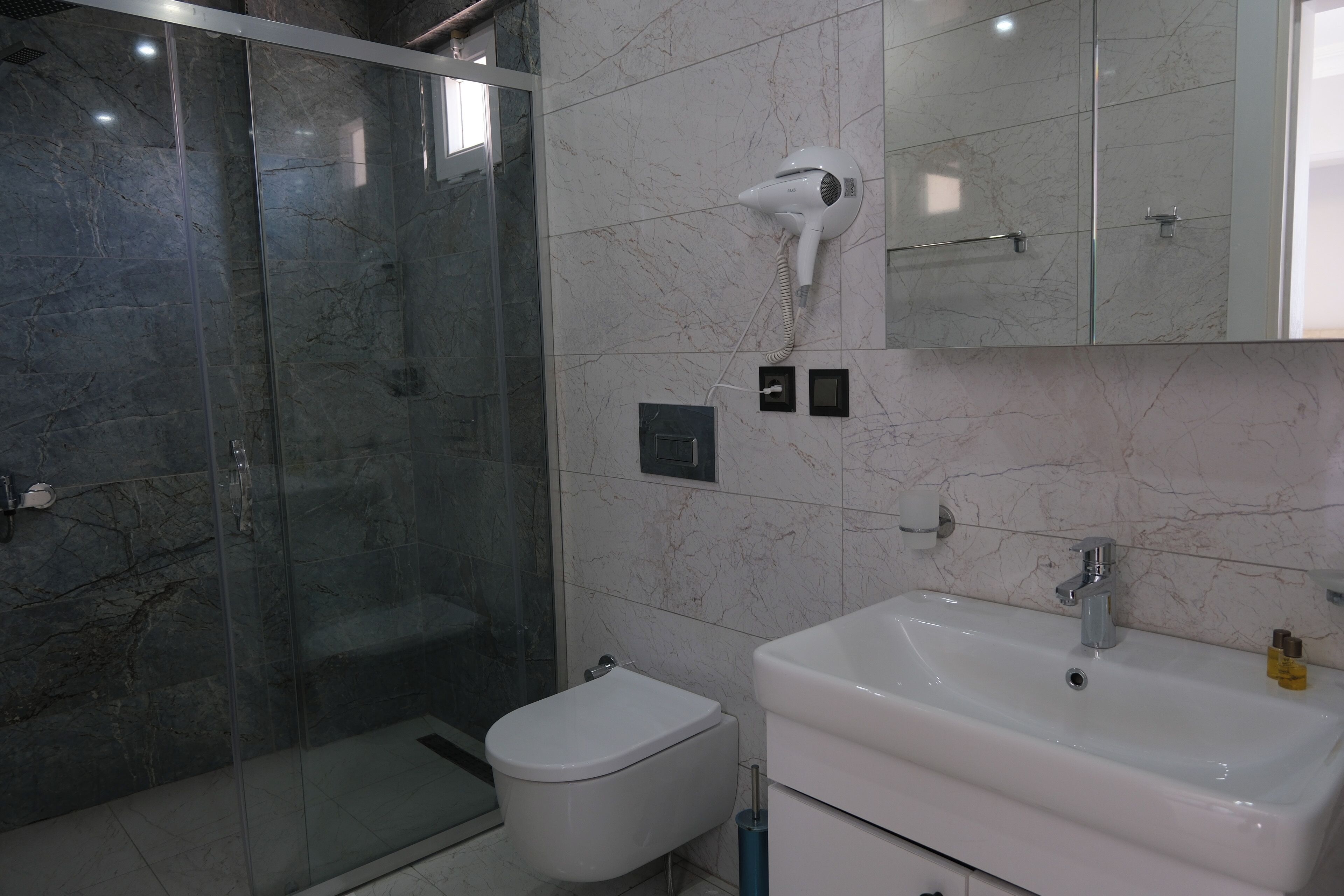 Une salle de bain avec une douche à parois de verre, des toilettes blanches, un lavabo et un sèche-cheveux fixé au mur.