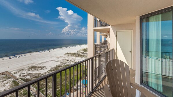 A balcony with a view of a beach, a pool, and a building with a glass door.