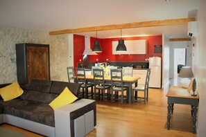 Un salon avec un canapé et des fauteuils, une table à manger avec des chaises, une cuisine à l’arrière-plan et une poutre en bois au plafond.