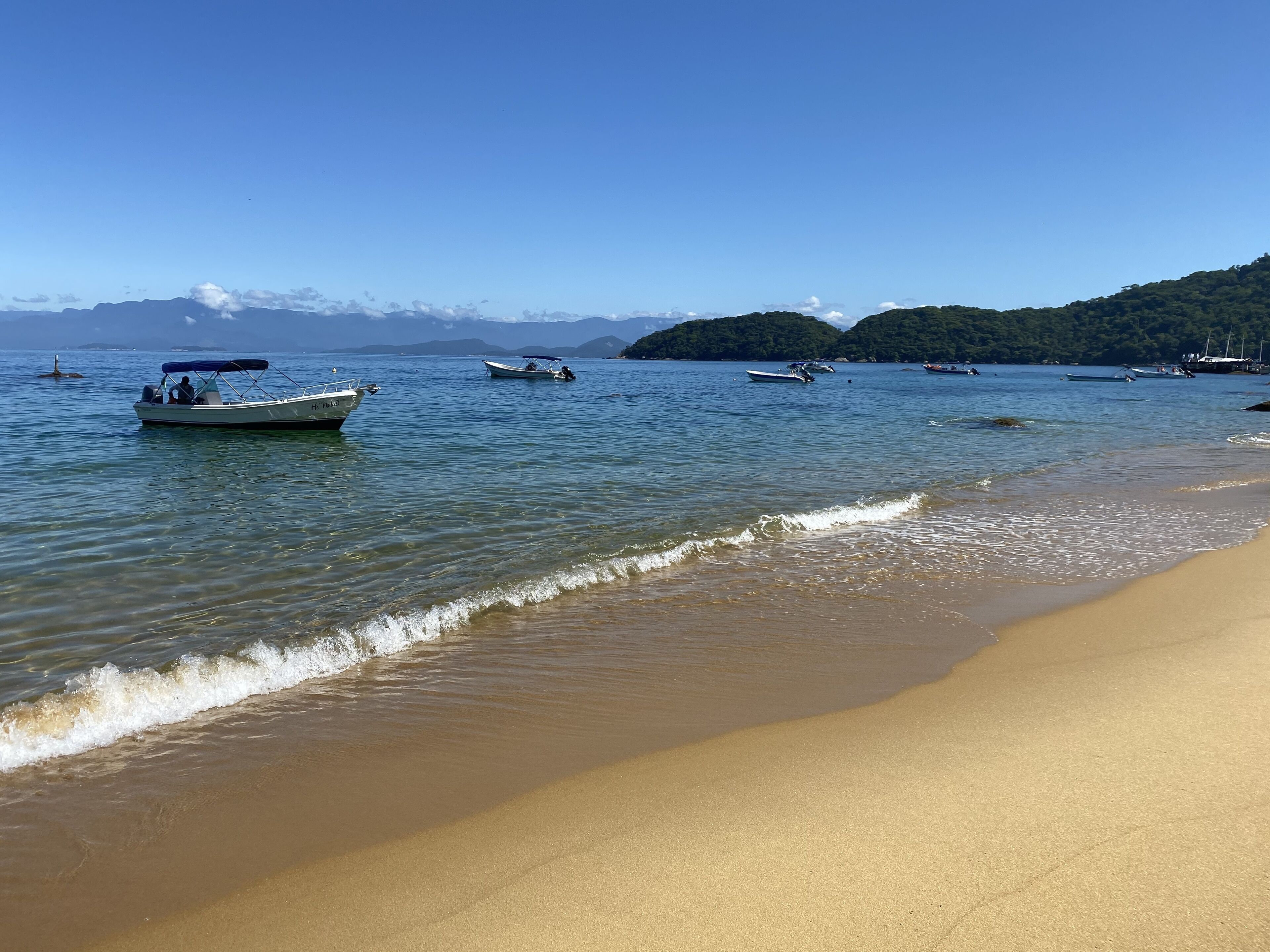 Una playa con agua cristalina, barcos y montañas a lo lejos.
