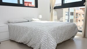 Een moderne slaapkamer met een groot bed, nachtkastje, raam met gordijnen en uitzicht op gebouwen.