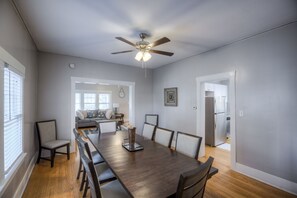 A dining area with a long wooden table, chairs, a ceiling fan, and a view into the living room. - Big Midtown Home! Sports Room, Bunk Beds! (Omaha)