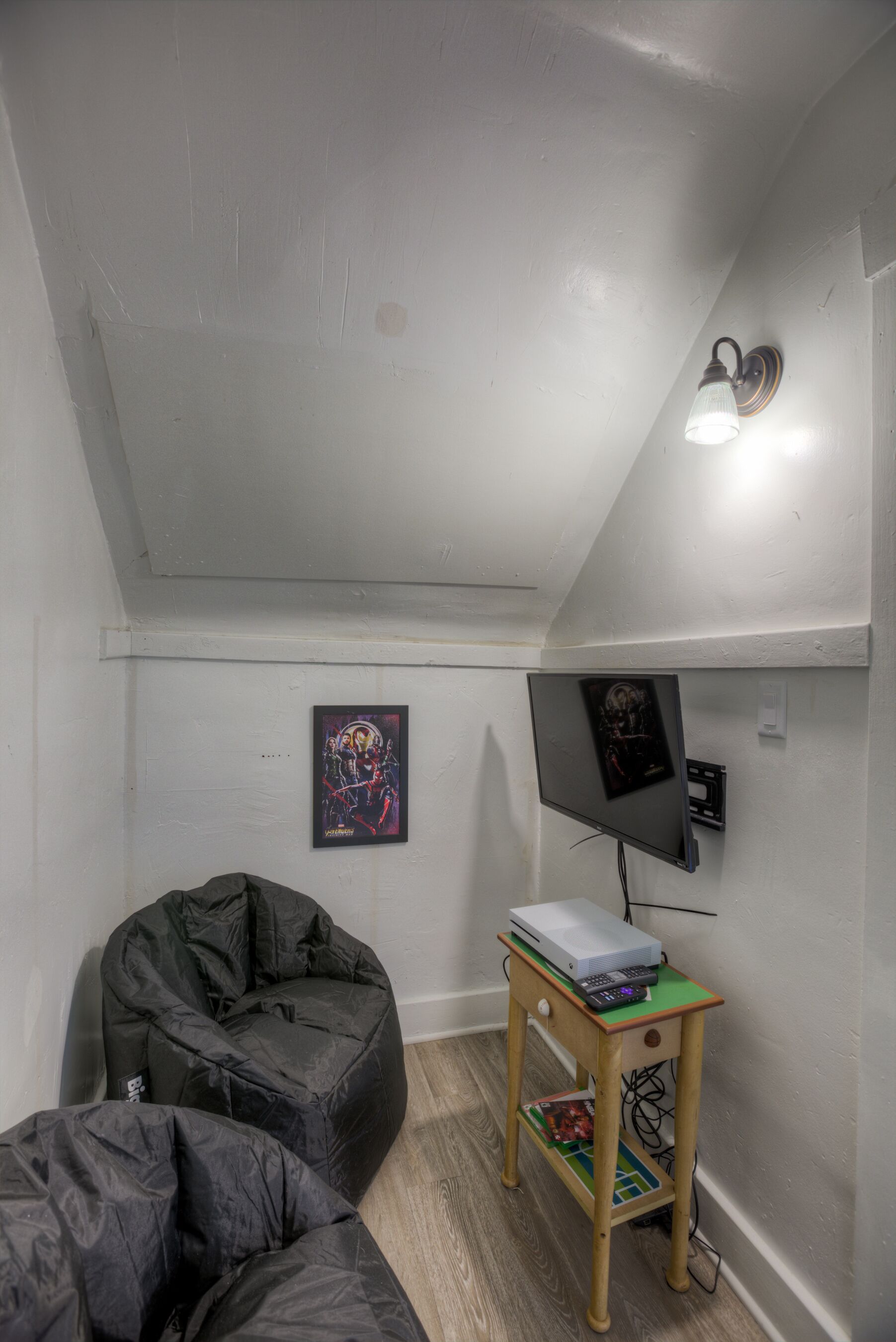 A small, white-walled room with a sloped ceiling, a black bean bag chair, a small wooden table with a TV and a projector, and a poster on the wall.