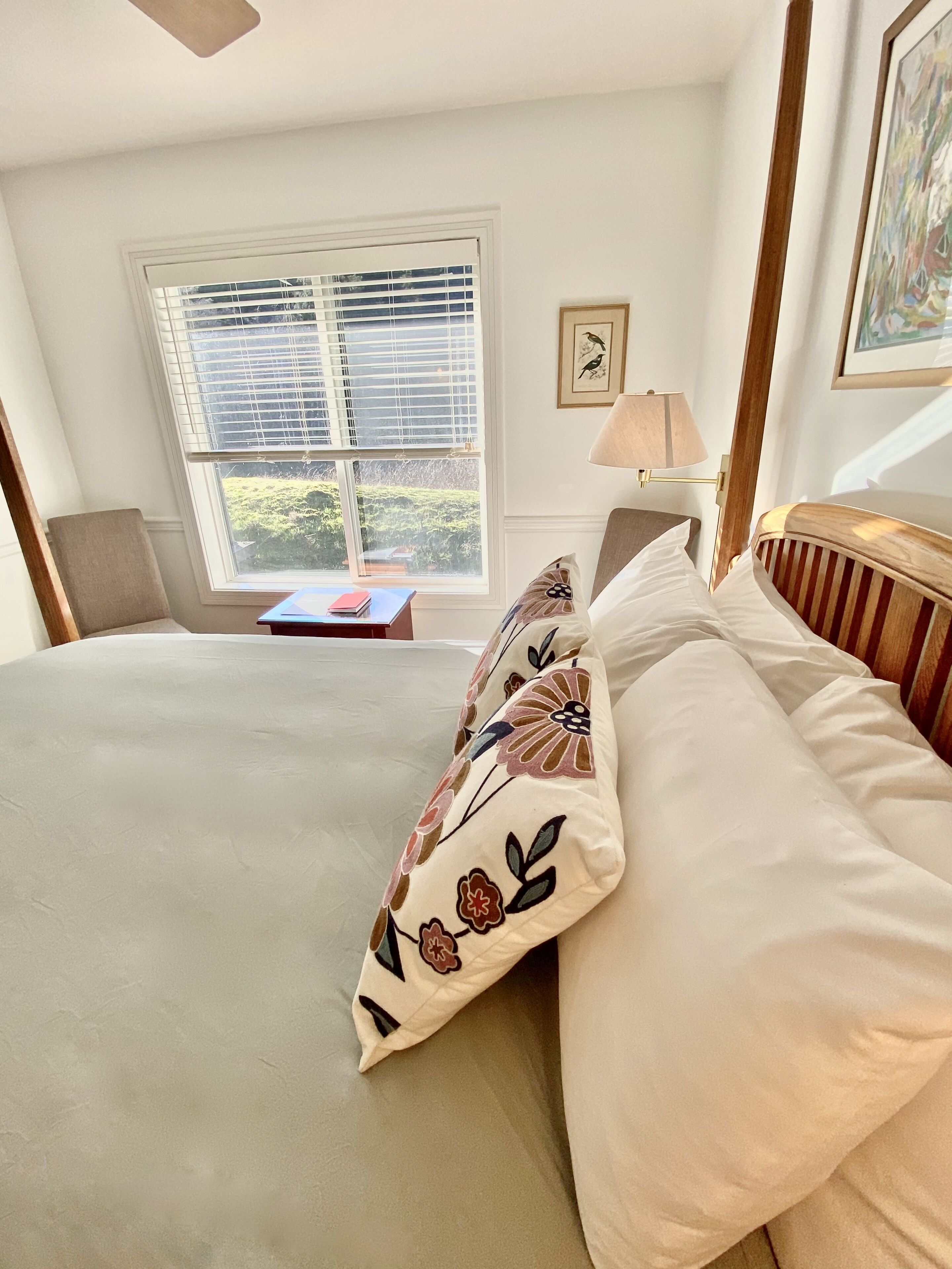 A bedroom with a bed, a chair, a table, a window with blinds, a lamp, and a framed picture on the wall.