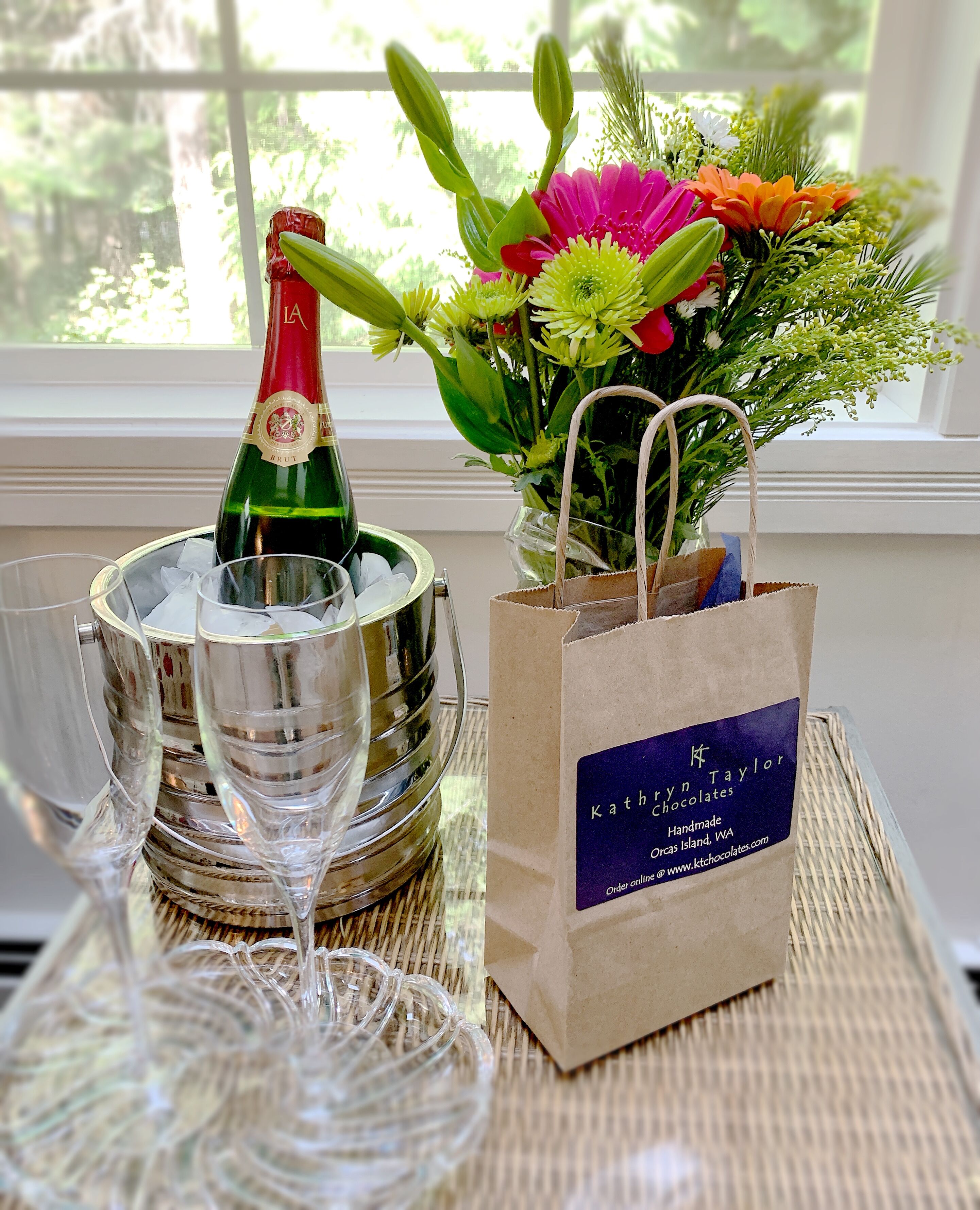 A bottle of champagne, two glasses, and a floral arrangement on a table near a window.