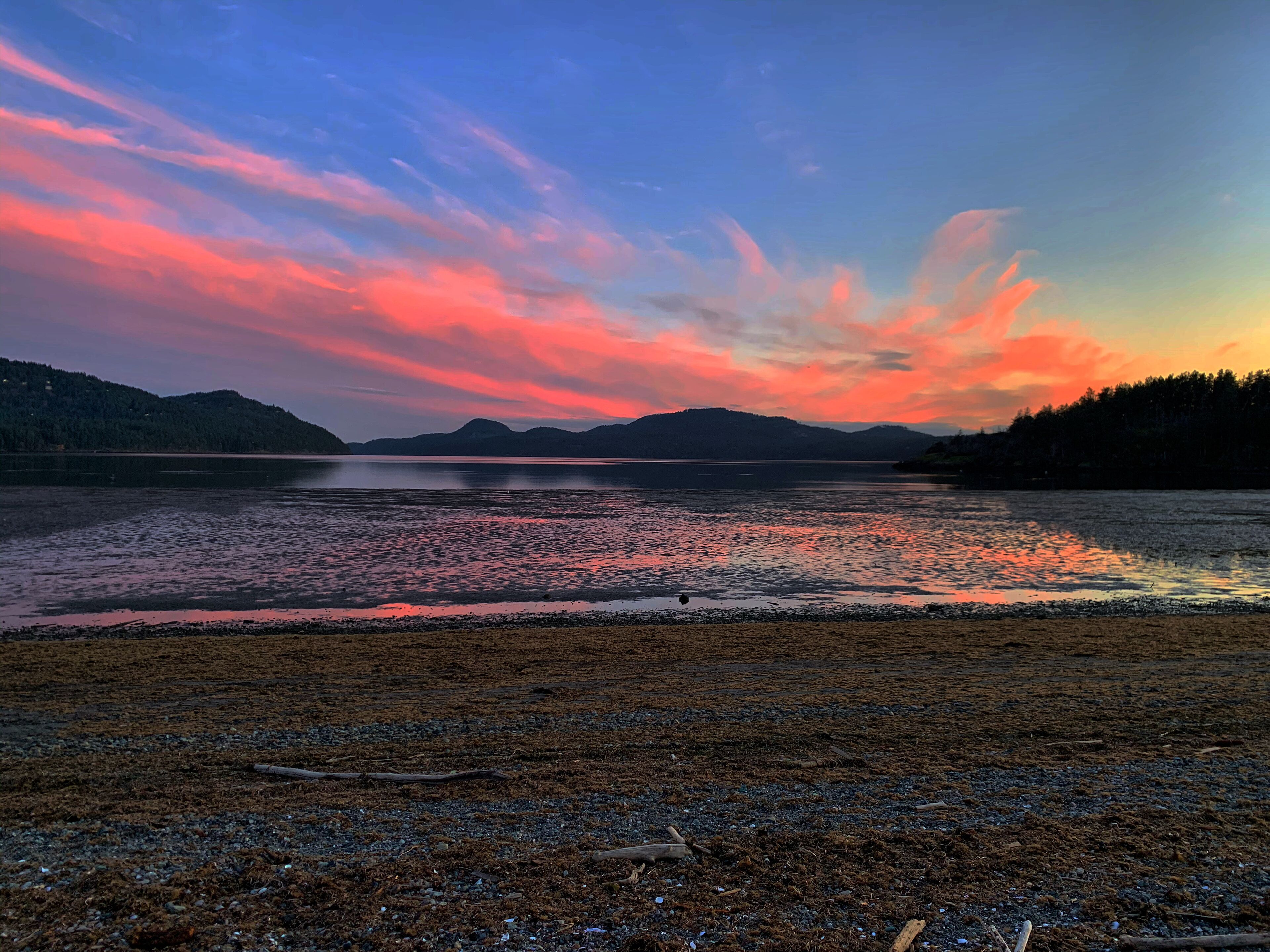 Een strand met een kalm meer, kiezelstenen en verre bergen onder een kleurrijke zonsondergang.