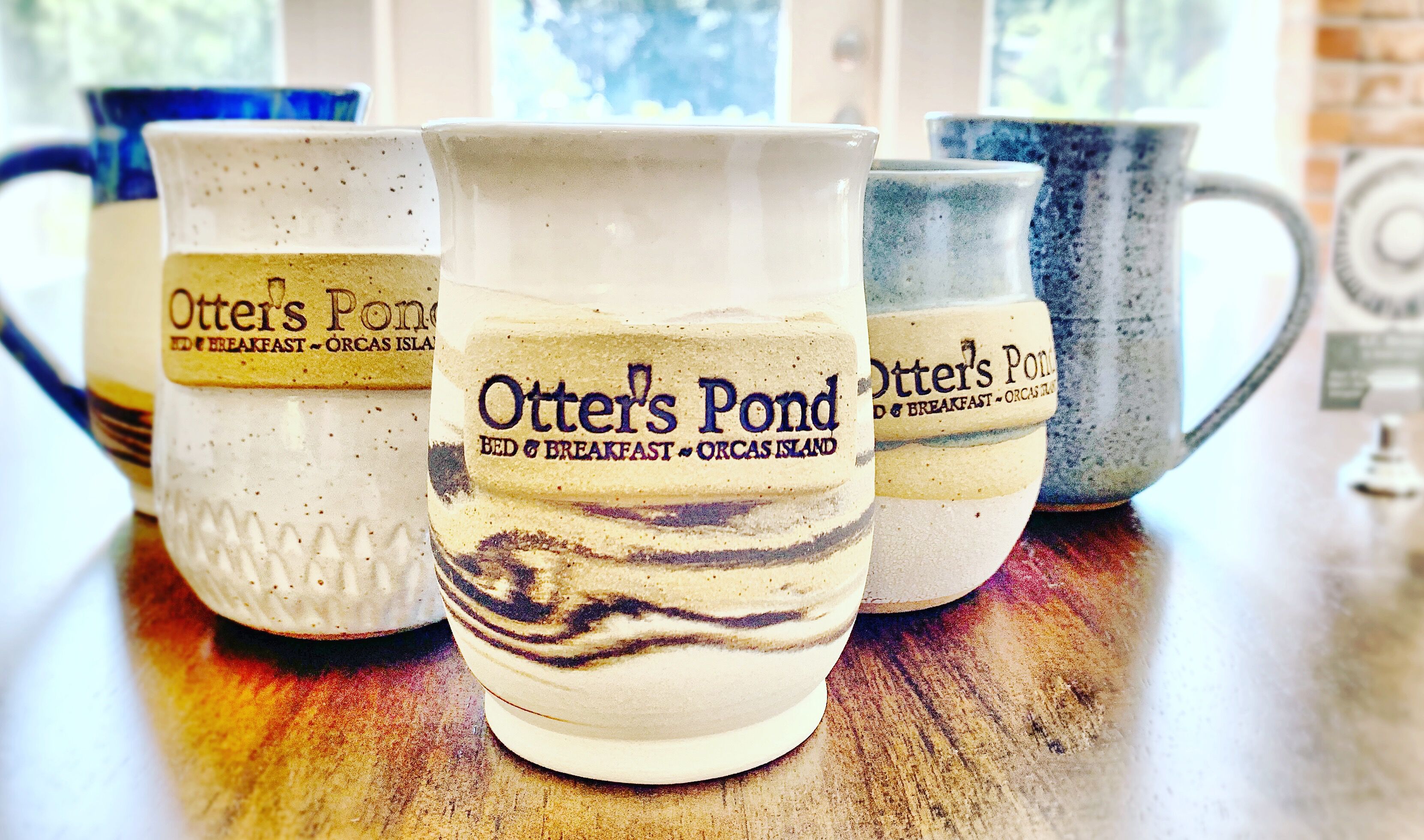 A row of ceramic mugs with the text "Otter's Pond Bed & Breakfast - Orcas Island" on them.