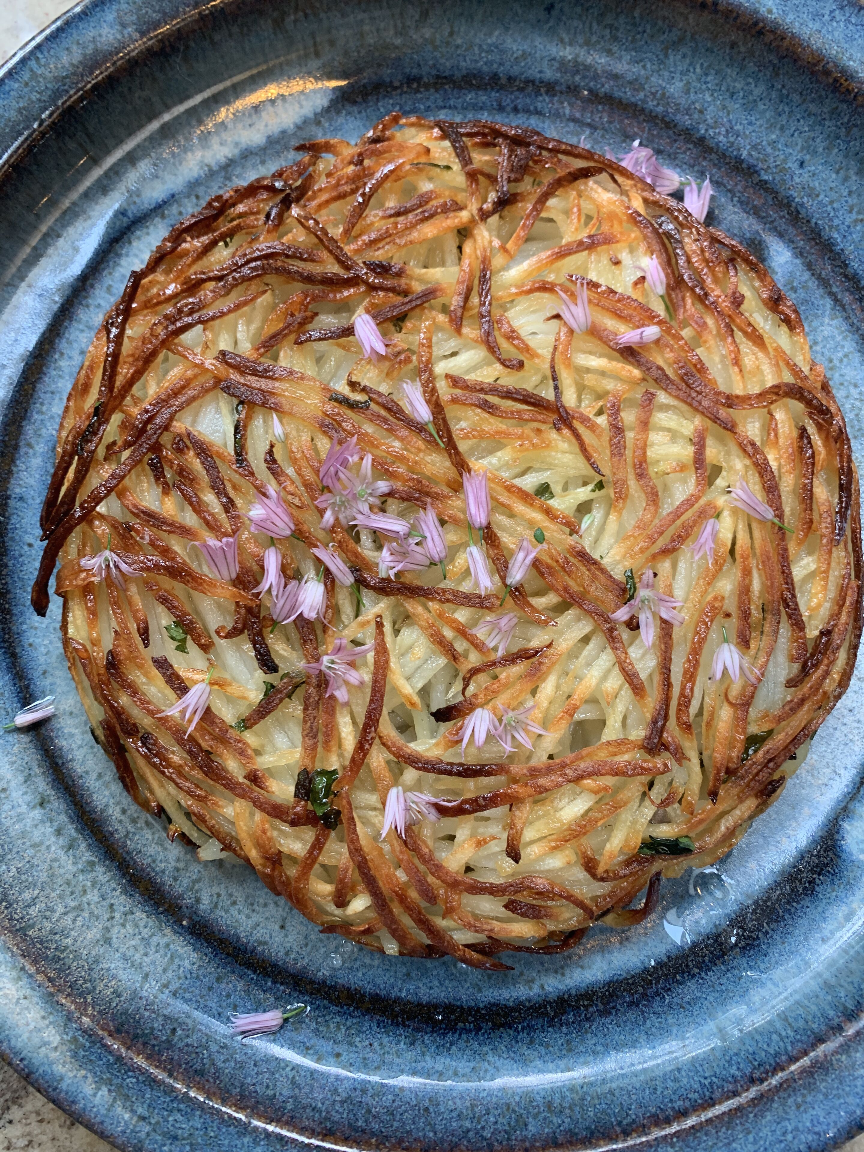 A round, golden-brown potato pancake with thin strips and small purple flowers on a blue ceramic plate.