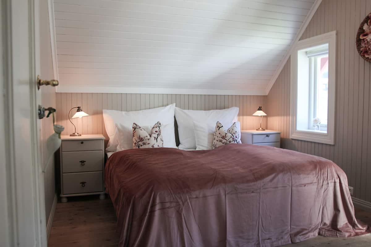 A bedroom with a sloped ceiling, a bed with a brown bedspread, two bedside tables with lamps, and a window with a view.