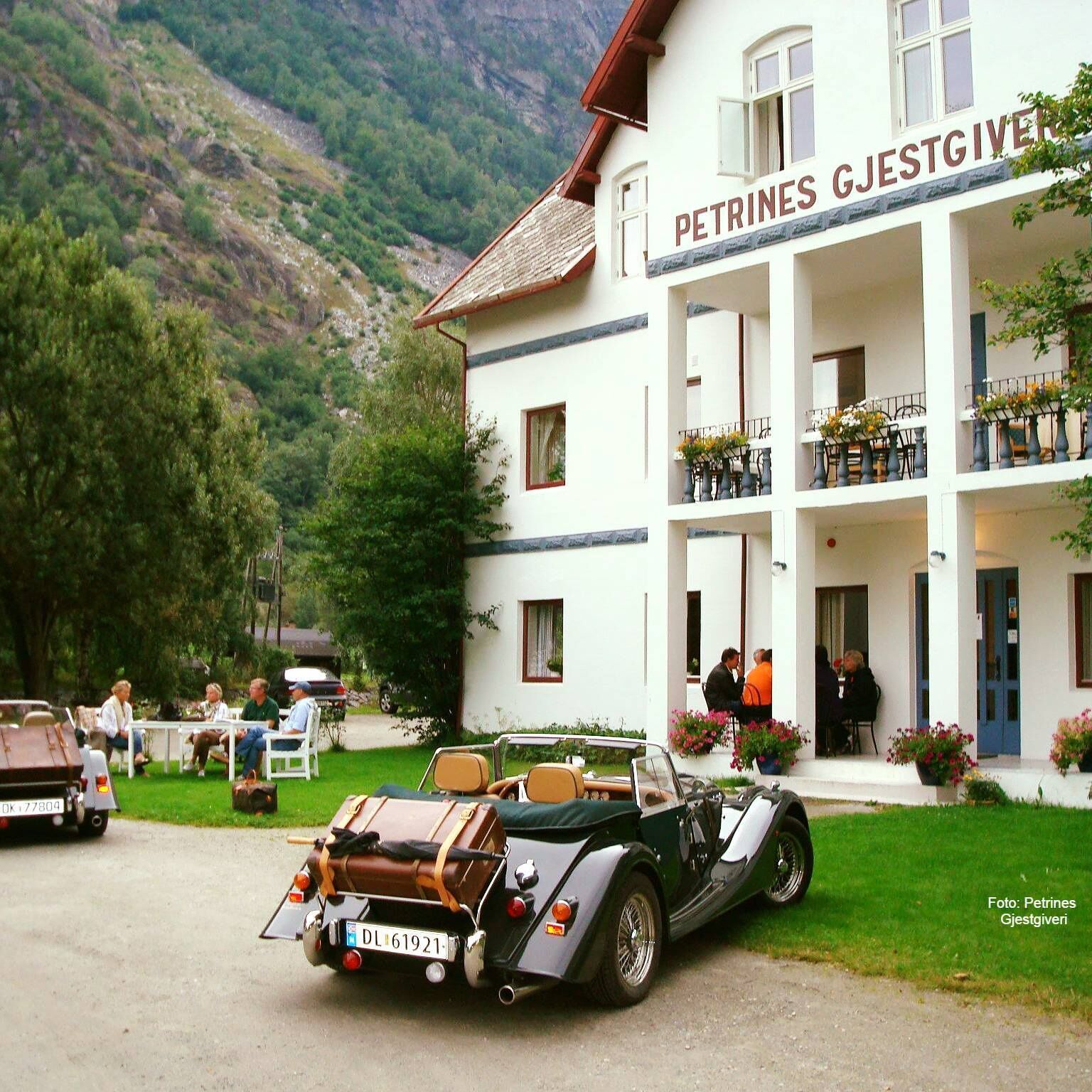 A two-story building with a sign reading "PETRINES GJESTGIVER". There are outdoor seating areas with tables and chairs, and a vintage car parked in front.