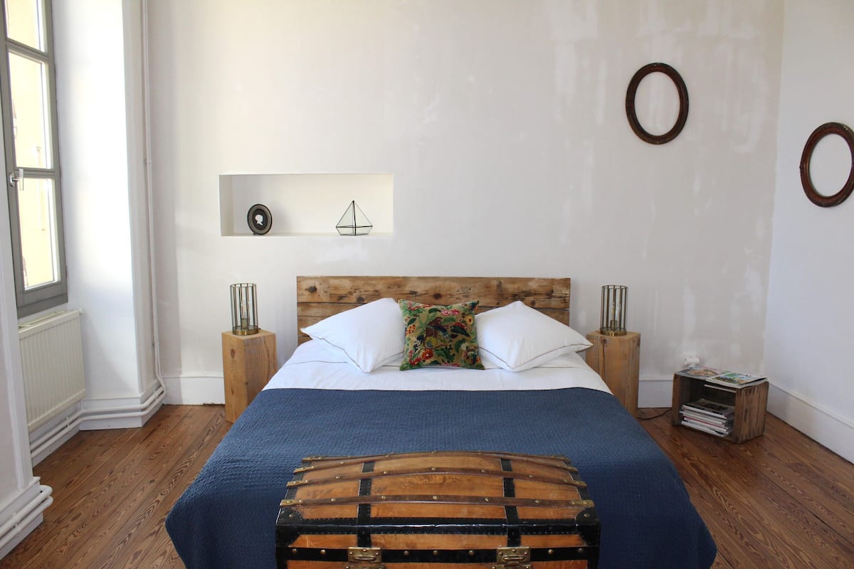 A bedroom with a wooden headboard, a bed with white linens, a blue blanket, a decorative trunk, and two bedside tables with lamps.