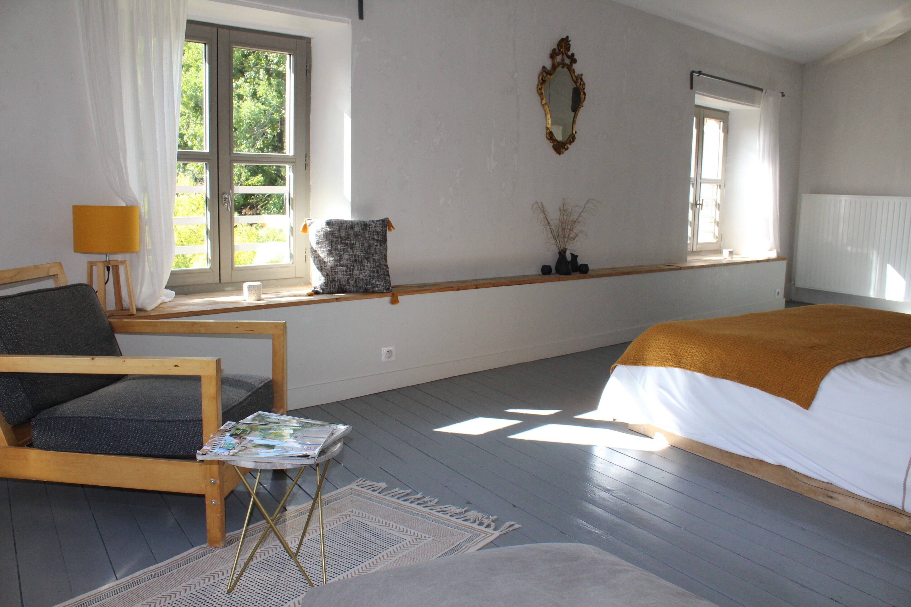 A bedroom with a bed, a chair, a small table with a magazine, a window with curtains, and a mirror on the wall.