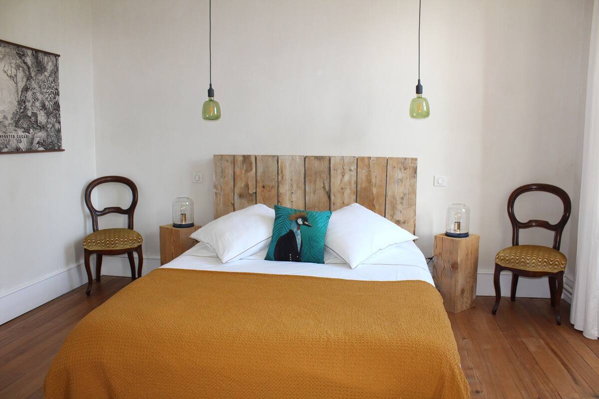 A bedroom with a wooden headboard, a large bed with a mustard-colored blanket, two wooden nightstands with lamps, and two upholstered chairs.