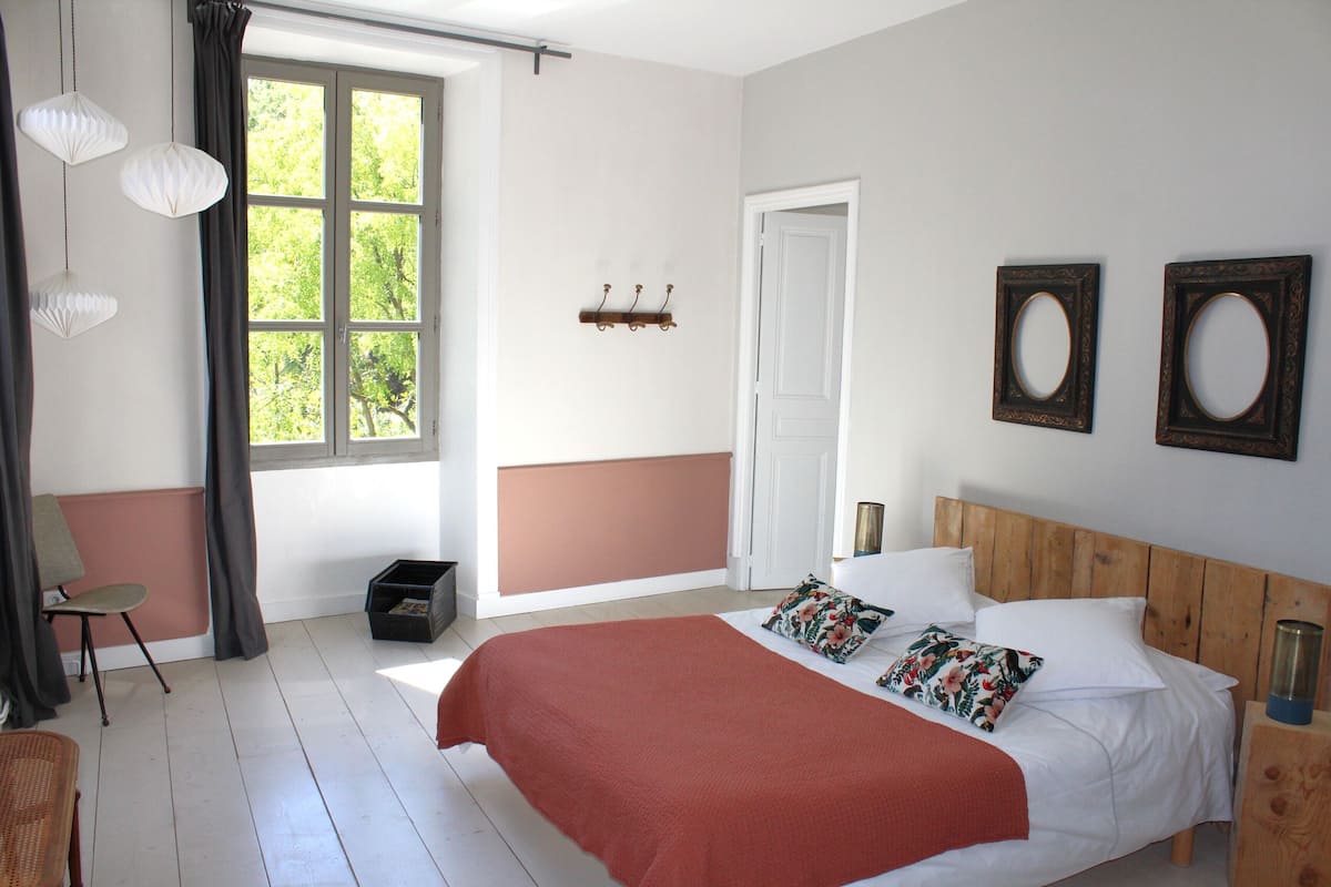 A bedroom with a large bed, a window with curtains, a wooden headboard, and a wall-mounted shelf with hooks.