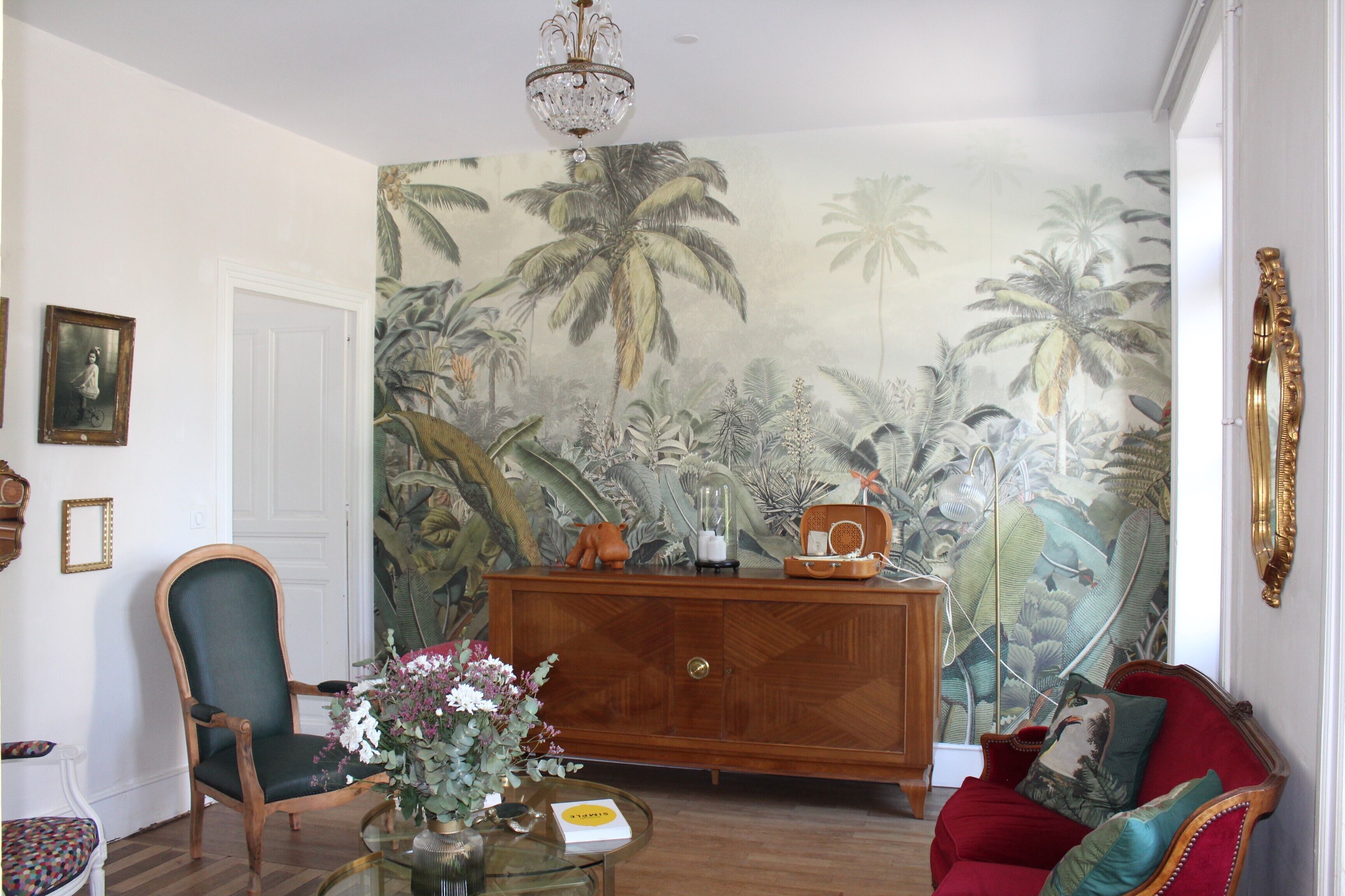 A room with a wooden cabinet, a red sofa, a green chair, and a wall with a tropical mural.