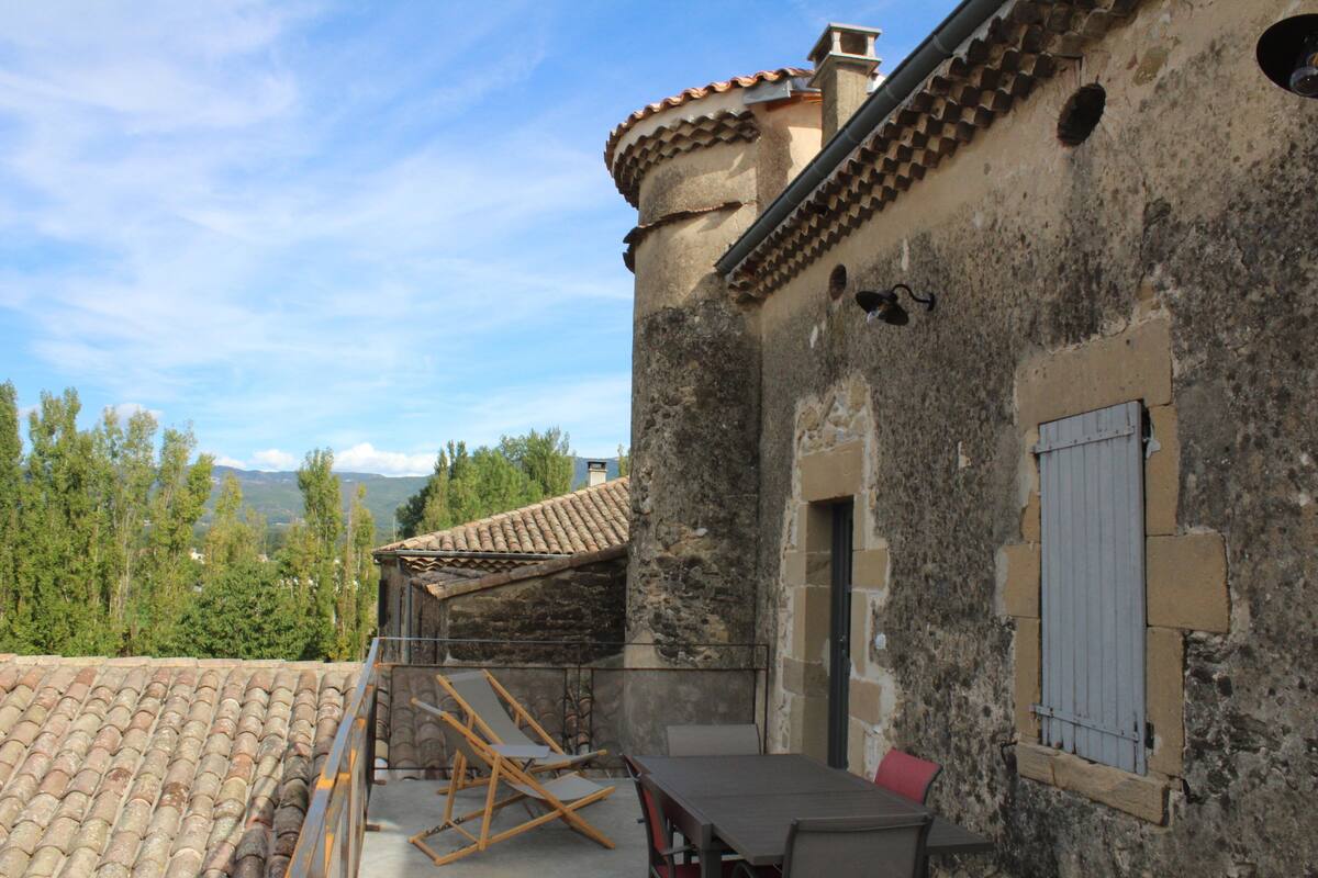 A rustic stone building with a tiled roof, a wooden table, and a red chair.